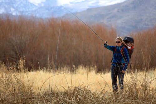 A person wearing sunglasses, a blue jacket, and dark pants fishes in a grassy area near Bishop, California with a child in a backpack carrier. Tall, dry grass and shrubs surround them, with mountains and snow in the background. The person holds a fishing rod cast toward the left. visit bishop