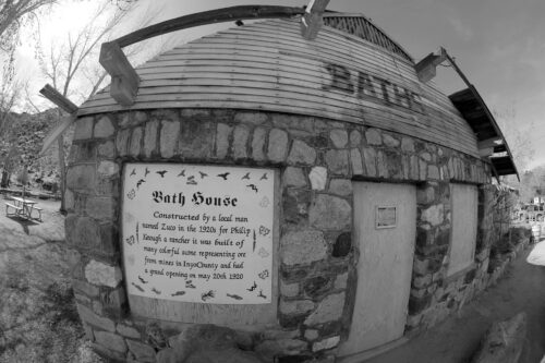 A black and white photo of a round stone building labeled "Bath House" in Bishop, California. A sign on the building reads: "Constructed by a local man named Zua in 1906 for Philip Knight's mother. Represents 20 cultures. Healed 5 good springs on May 26th, 1906." A door is adjacent. visit bishop