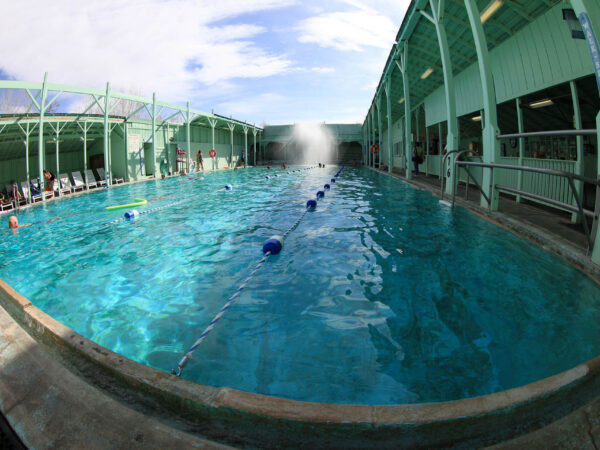 An indoor swimming pool in Bishop, California boasts clear blue water, divided into lanes by blue and white floating markers. The pool is surrounded by a green-painted structure, with some people sitting and standing at the sides. The ceiling is partially open, revealing a blue sky. visit bishop