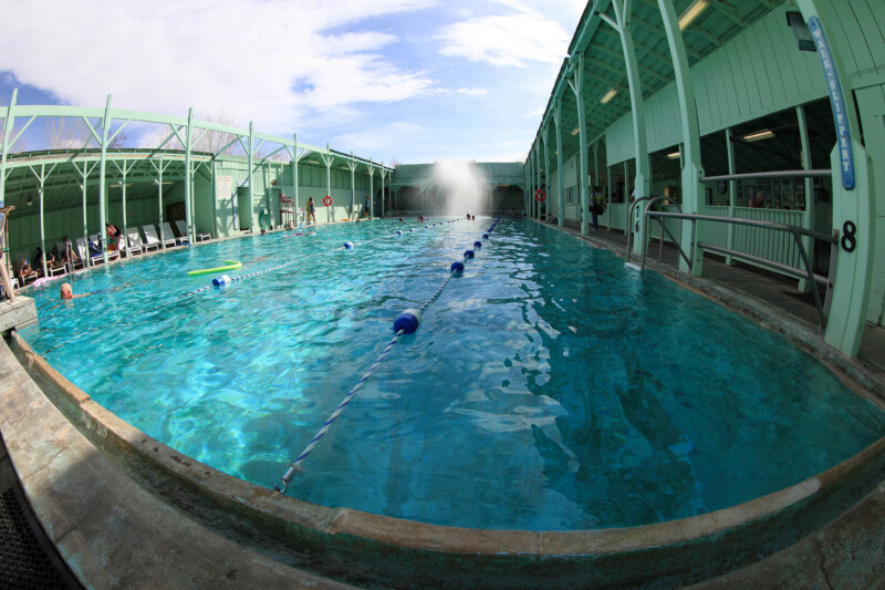 An indoor swimming pool in Bishop, California boasts clear blue water, divided into lanes by blue and white floating markers. The pool is surrounded by a green-painted structure, with some people sitting and standing at the sides. The ceiling is partially open, revealing a blue sky. visit bishop