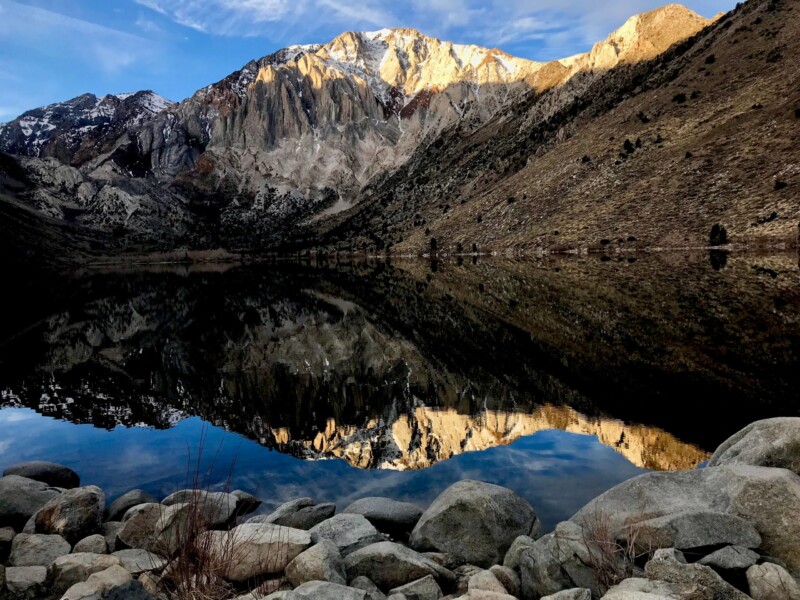 A serene mountain landscape in Bishop, California, with rocky peaks and slopes reflected perfectly in a calm lake. The foreground features large rocks along the lake's edge, and the sky is clear with a few clouds. The sunlight casts a golden hue on the mountain tops. visit bishop