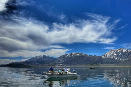 A small motorboat with three people is floating on a calm, reflective lake near Bishop, California. Snow-capped mountains can be seen in the background, and the sky is partly cloudy with some dramatic cloud formations. Another boat is visible in the distance. visit bishop