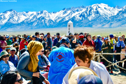 A diverse crowd gathers near a monument with snow-capped mountains in the background on a sunny day. visit bishop