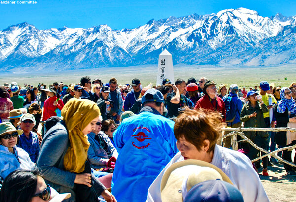 A diverse crowd gathers near a monument with snow-capped mountains in the background on a sunny day. visit bishop