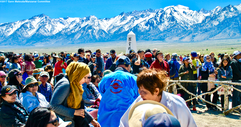 A diverse crowd gathers near a monument with snow-capped mountains in the background on a sunny day. visit bishop