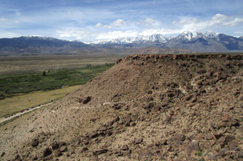 A rocky hilltop in Bishop, California, offers a breathtaking view of a vast green valley, with snow-capped mountains in the distance under a partly cloudy sky. Sparse vegetation dots the hill while lush greenery thrives below. Dirt paths are visible leading into the serene valley. visit bishop