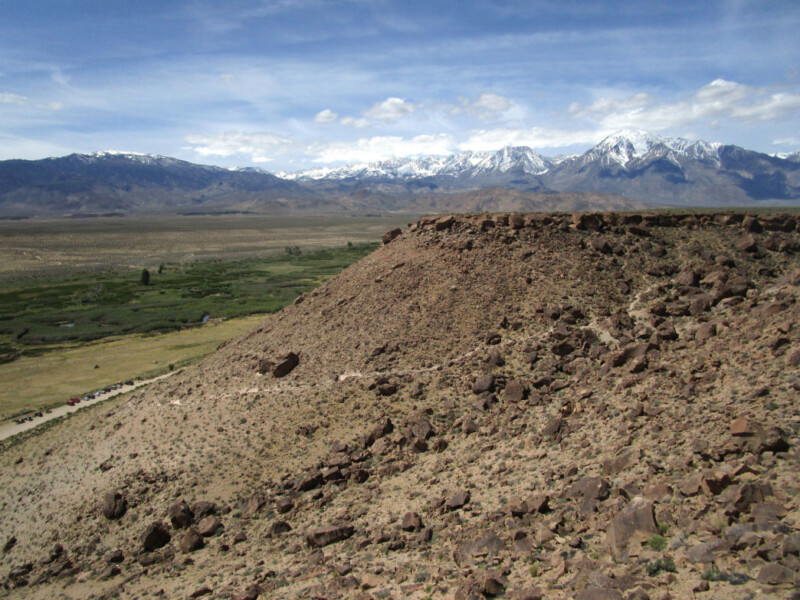 A rocky hilltop in Bishop, California, offers a breathtaking view of a vast green valley, with snow-capped mountains in the distance under a partly cloudy sky. Sparse vegetation dots the hill while lush greenery thrives below. Dirt paths are visible leading into the serene valley. visit bishop