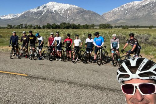 A group of cyclists pose for a photo on a scenic road near Bishop, California, with snow-capped mountains in the background. visit bishop