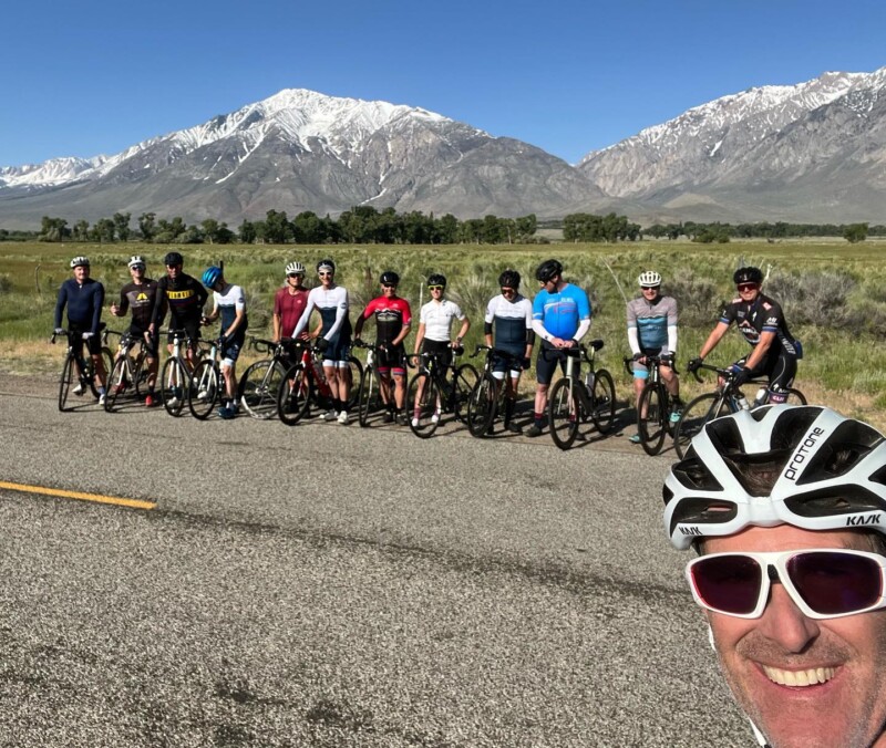 A group of cyclists pose for a photo on a scenic road near Bishop, California, with snow-capped mountains in the background. visit bishop