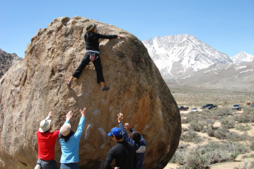 A person boulders up a large rock as four spotters extend their hands towards them in the stunning, mountainous landscape of Bishop, California. visit bishop