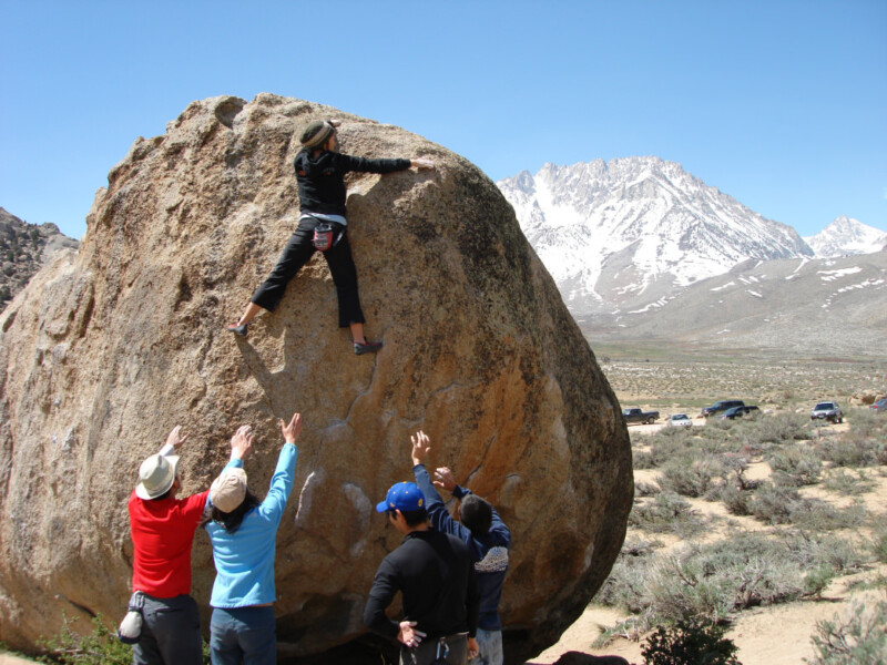 A person boulders up a large rock as four spotters extend their hands towards them in the stunning, mountainous landscape of Bishop, California. visit bishop