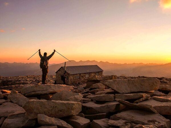 A hiker stands triumphantly with arms raised, holding trekking poles, on a rocky mountain summit at sunset near Bishop, California. A small stone cabin is visible in the background, with layered mountain ridges extending into the distance under a colorful sky. visit bishop