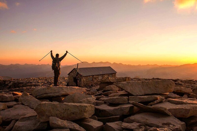 A hiker stands triumphantly with arms raised, holding trekking poles, on a rocky mountain summit at sunset near Bishop, California. A small stone cabin is visible in the background, with layered mountain ridges extending into the distance under a colorful sky. visit bishop