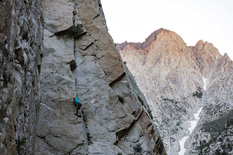 A rock climber scales a large, steep cliff with a rugged mountain landscape in the background. visit bishop