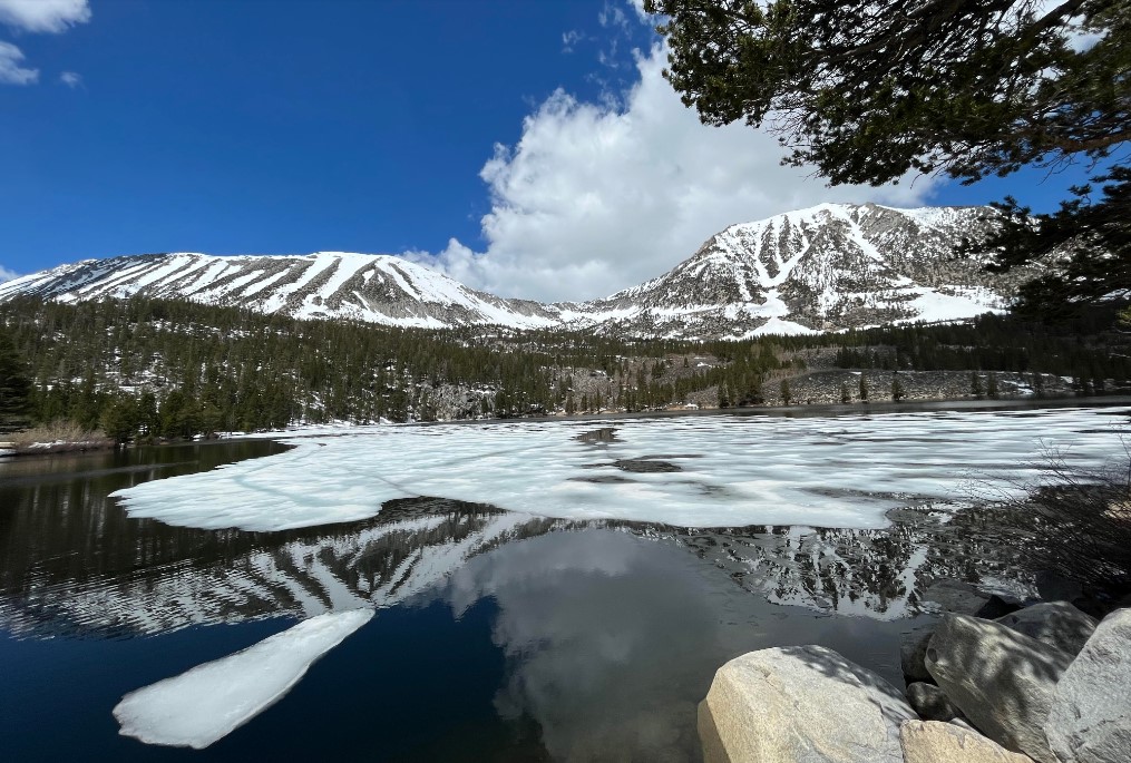 Lake partly covered with melting ice, reflecting snow-capped mountains and a clear blue sky with scattered clouds in the Eastern Sierra near Bishop, California. visit bishop