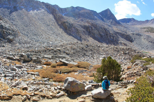 A lone hiker wearing blue sits on a large rock in front of the vast mountainous landscape near Bishop, California. The rocky slopes and sparse vegetation enhance the serene and remote wilderness setting under a clear sky with a few clouds. visit bishop