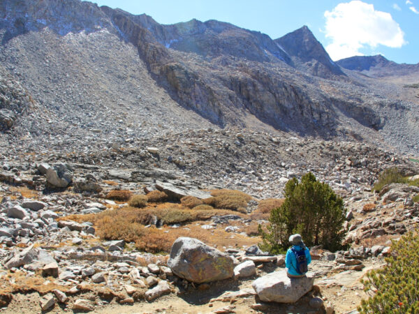 A lone hiker wearing blue sits on a large rock in front of the vast mountainous landscape near Bishop, California. The rocky slopes and sparse vegetation enhance the serene and remote wilderness setting under a clear sky with a few clouds. visit bishop
