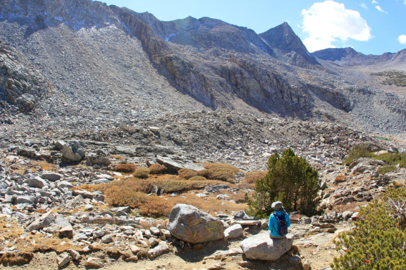 A lone hiker wearing blue sits on a large rock in front of the vast mountainous landscape near Bishop, California. The rocky slopes and sparse vegetation enhance the serene and remote wilderness setting under a clear sky with a few clouds. visit bishop