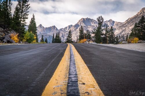 Close-up of a road with a yellow center line, leading to the mountains near Bishop, California, surrounded by trees under a cloudy sky. visit bishop