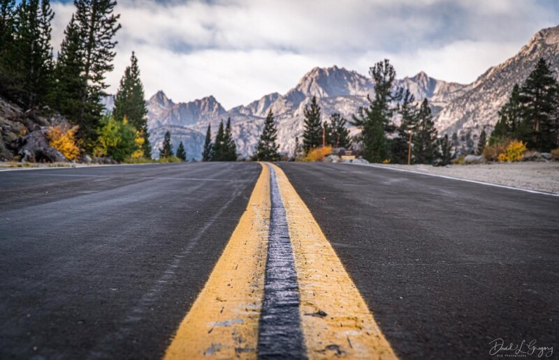 Close-up of a road with a yellow center line, leading to the mountains near Bishop, California, surrounded by trees under a cloudy sky. visit bishop