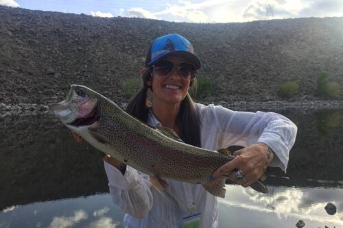 A person wearing a blue cap, sunglasses, and a white shirt is standing by a river in Bishop, California, proudly holding a large trout. The background features a rocky embankment and a partly cloudy sky reflecting on the water. visit bishop