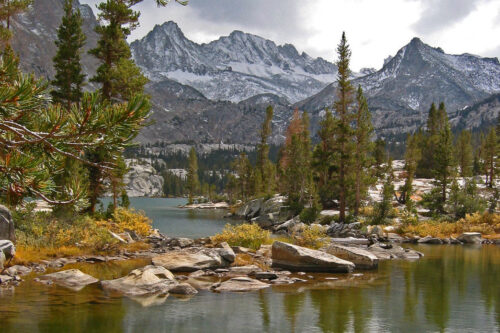 A serene lake near Bishop, California, surrounded by evergreen trees and rocky terrain under a cloudy sky. Snow-capped mountains rise in the distance, reflecting in the calm water. The scene exudes a peaceful, natural beauty with hints of autumn colors among the foliage. visit bishop