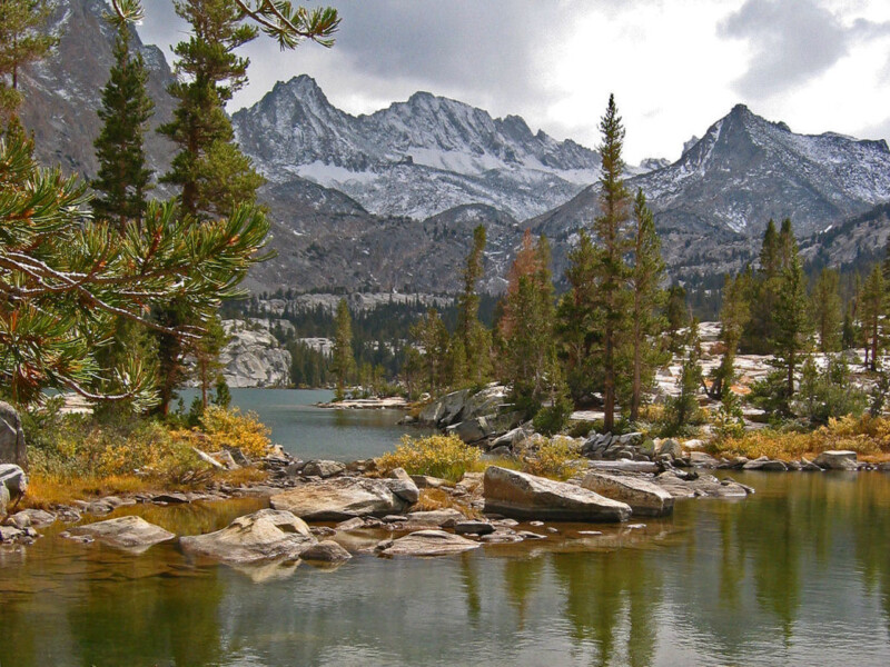 A serene lake near Bishop, California, surrounded by evergreen trees and rocky terrain under a cloudy sky. Snow-capped mountains rise in the distance, reflecting in the calm water. The scene exudes a peaceful, natural beauty with hints of autumn colors among the foliage. visit bishop