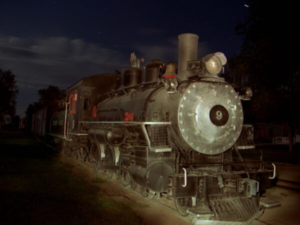 Old steam locomotive under a starry night sky, surrounded by trees and faintly visible buildings in the background. visit bishop