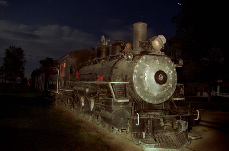 Old steam locomotive under a starry night sky, surrounded by trees and faintly visible buildings in the background. visit bishop