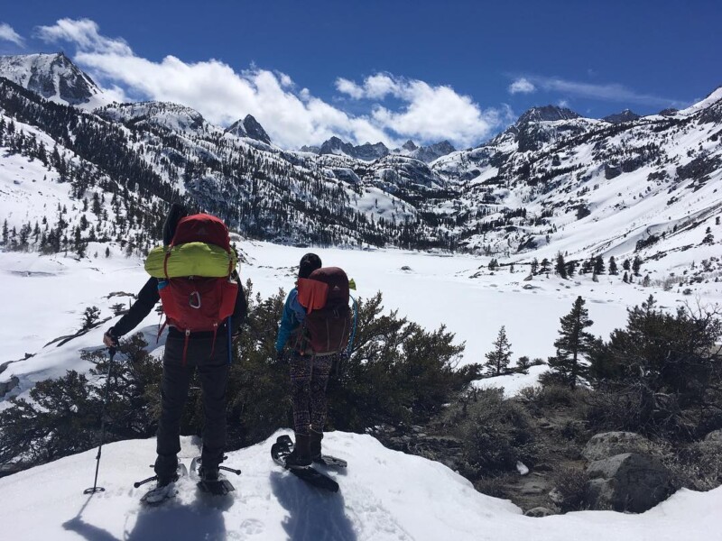 Two hikers with backpacks stand on a snowy mountain trail, overlooking a vast landscape of snowy peaks and valleys. visit bishop