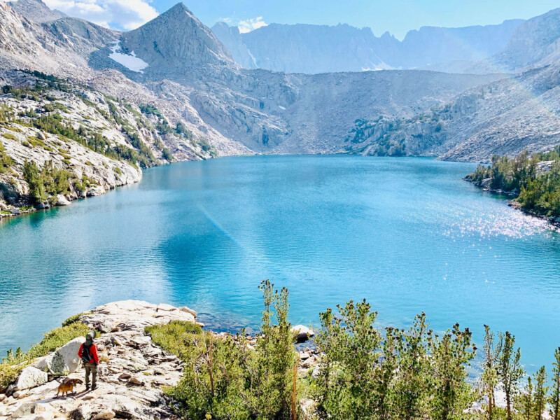 A hiker wearing a red jacket stands on a rocky ledge next to a dog, overlooking a large, clear blue mountain lake surrounded by rocky cliffs and evergreen trees in Bishop, California. The sun shines brightly, creating sparkling reflections on the water. Snow-capped peaks are in the distance. visit bishop