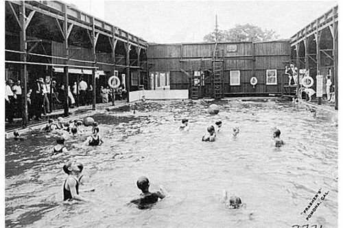 Black and white photo of a crowded indoor swimming pool in Bishop, California, with several people swimming and playing. Wood-paneled walls and a second-floor balcony enclose the pool area. A sign reads "SPAPERA'S" and a ladder leads to a diving platform. Spectators are seen on the left. visit bishop