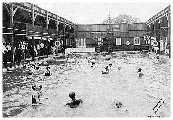 Black and white photo of a crowded indoor swimming pool in Bishop, California, with several people swimming and playing. Wood-paneled walls and a second-floor balcony enclose the pool area. A sign reads "SPAPERA'S" and a ladder leads to a diving platform. Spectators are seen on the left. visit bishop