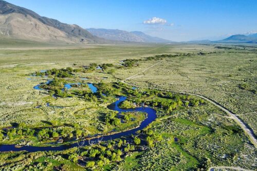 Aerial view of a winding river flowing through a green valley with mountains in the background under a clear blue sky. visit bishop