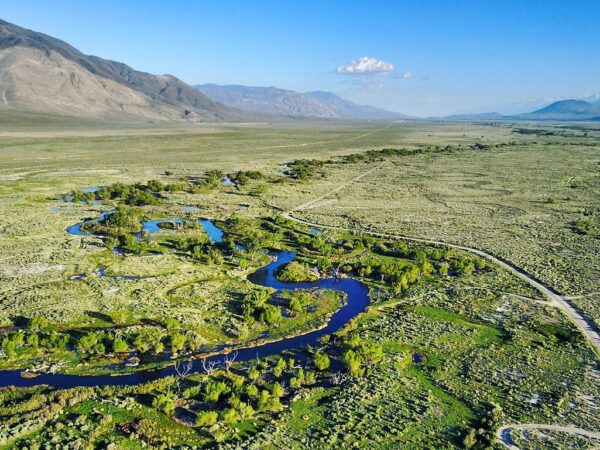 Aerial view of a winding river flowing through a green valley with mountains in the background under a clear blue sky. visit bishop