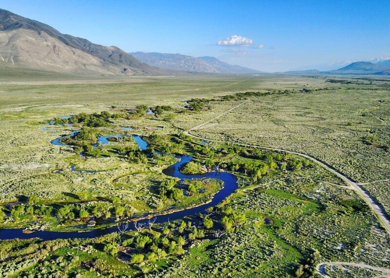 Aerial view of a winding river flowing through a green valley with mountains in the background under a clear blue sky. visit bishop