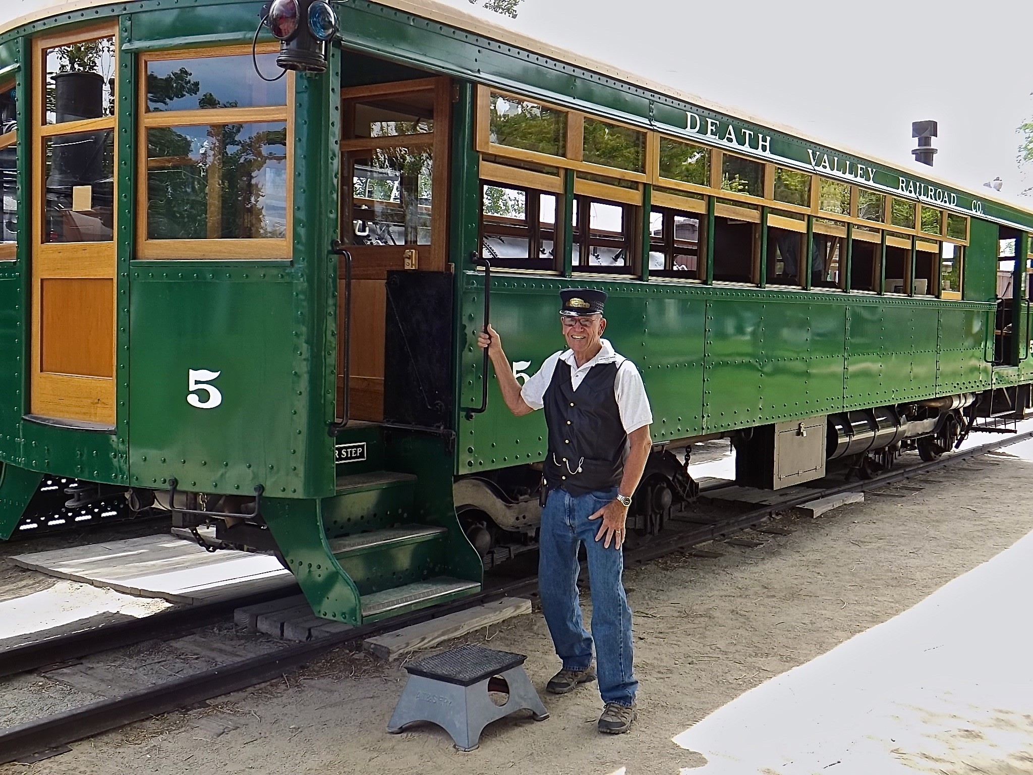 A man in uniform stands next to a green historic tram numbered 5 labeled "Death Valley Railroad," an evocative symbol of the rich history of Eastern Sierra. visit bishop