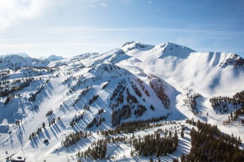 A vast snow-covered mountain range near Bishop, California, boasts peaks and ridges under a clear blue sky. Pine trees are scattered across the slopes, with small structures visible in the lower left corner, likely parts of a ski resort. Ski trails are also noticeable along the slopes. visit bishop