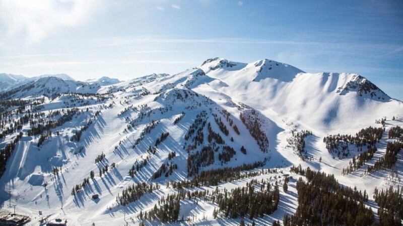 A vast snow-covered mountain range near Bishop, California, boasts peaks and ridges under a clear blue sky. Pine trees are scattered across the slopes, with small structures visible in the lower left corner, likely parts of a ski resort. Ski trails are also noticeable along the slopes. visit bishop