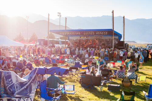 A crowd at an outdoor music concert in Bishop, California, with the majestic Eastern Sierra mountains in the background and chairs covering the grassy field. visit bishop