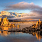 A serene lake with rocky formations reflecting on the water's surface, backdropped by snow-capped mountains under a sky with scattered clouds in Bishop, California. The golden light of the setting or rising sun illuminates the landscape, creating a warm, tranquil scene. visit bishop