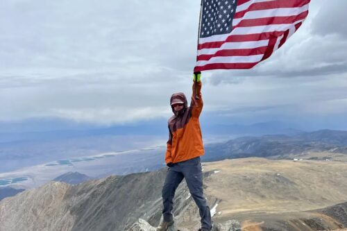 Person in hiking attire stands atop a rocky mountain summit, holding an American flag, with vast landscape in the background. visit bishop