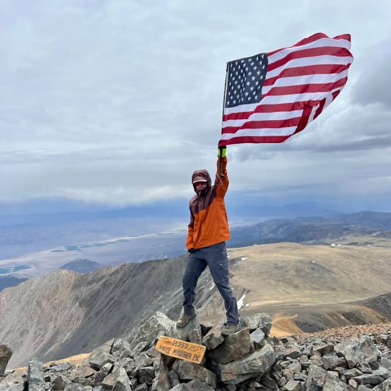 Person in hiking attire stands atop a rocky mountain summit, holding an American flag, with vast landscape in the background. visit bishop