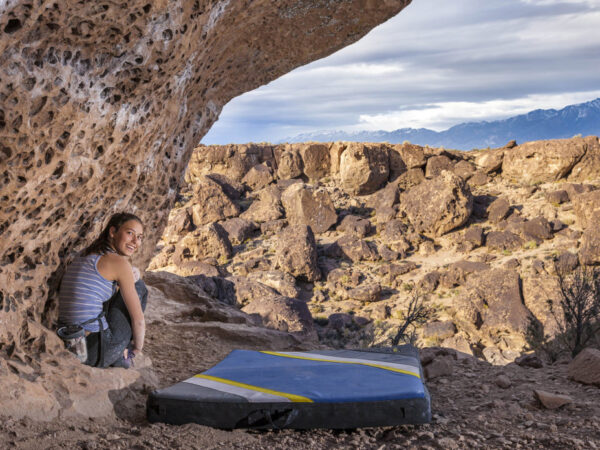 A woman sits under a large, rock overhang in the rocky desert landscape of Bishop, California, smiling at the camera. She is dressed in athletic gear with a climbing harness and a crash pad placed on the ground beside her. Mountains are visible in the distance under a cloudy sky. visit bishop