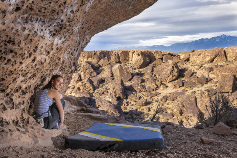 A woman sits under a large, rock overhang in the rocky desert landscape of Bishop, California, smiling at the camera. She is dressed in athletic gear with a climbing harness and a crash pad placed on the ground beside her. Mountains are visible in the distance under a cloudy sky. visit bishop