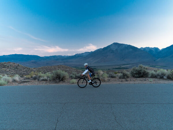 A person in cycling gear rides a bicycle on an empty road through the desert landscape of Bishop, California, at dusk. Mountains and an expansive sky with hues of blue and pink serve as the backdrop, while scrub vegetation lines the roadside. visit bishop