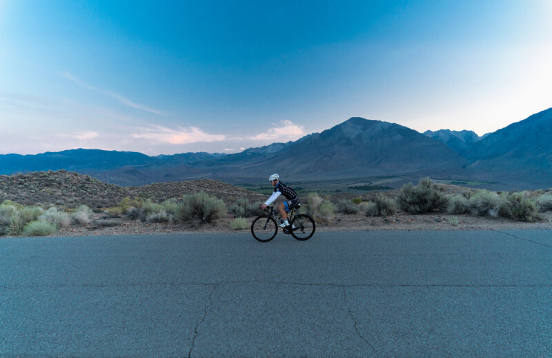 A person in cycling gear rides a bicycle on an empty road through the desert landscape of Bishop, California, at dusk. Mountains and an expansive sky with hues of blue and pink serve as the backdrop, while scrub vegetation lines the roadside. visit bishop