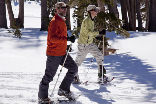 Two people snowshoeing in a snowy forest, wearing warm clothing and sunglasses, with trees and snow visible in the background. visit bishop