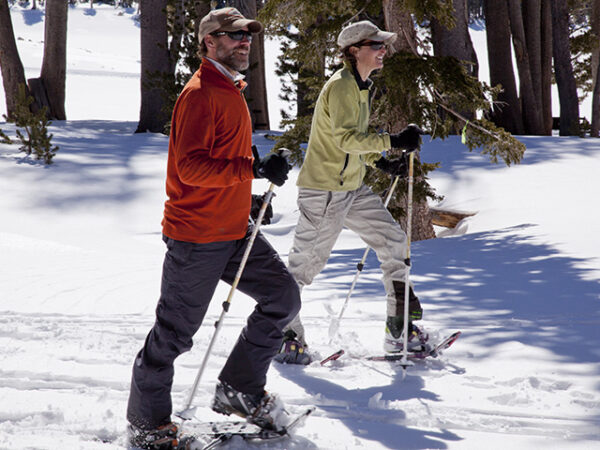 Two people snowshoeing in a snowy forest, wearing warm clothing and sunglasses, with trees and snow visible in the background. visit bishop