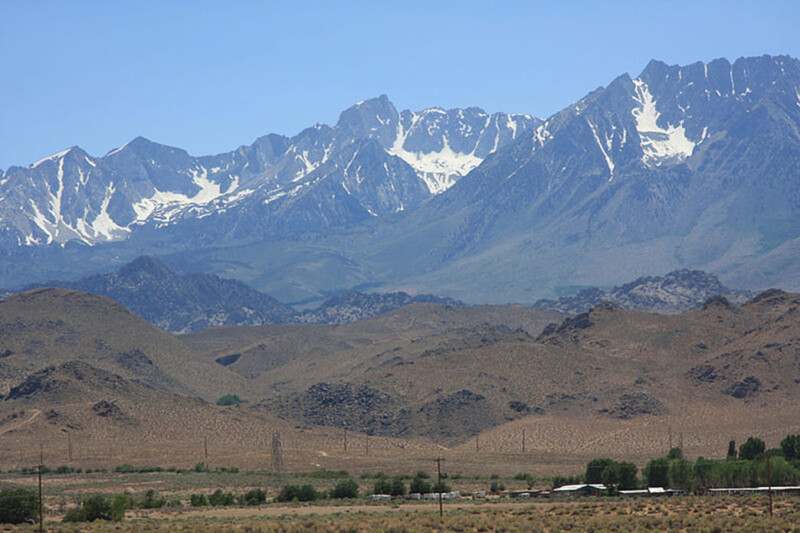 A rugged mountain range with snow-capped peaks stands against a clear blue sky. The foreground features arid, rocky terrain with sparse vegetation and a few small buildings, reminiscent of the breathtaking landscapes near Bishop, California. visit bishop